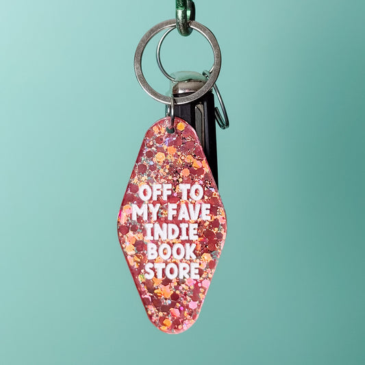 A glittery pink and orange keychain with white text reading 'OFF TO MY FAVE INDIE BOOK STORE' hangs from a green carabiner on a wooden rod against a light teal background.