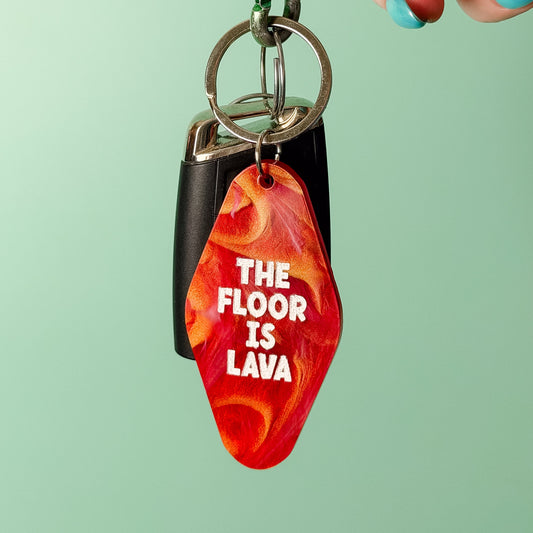A hand with teal nail polish holds a keychain featuring a red and orange lava-patterned tag with the text 'THE FLOOR IS LAVA' in white. The keychain also includes a black rectangular tag and silver metal rings, set against a solid mint green background.