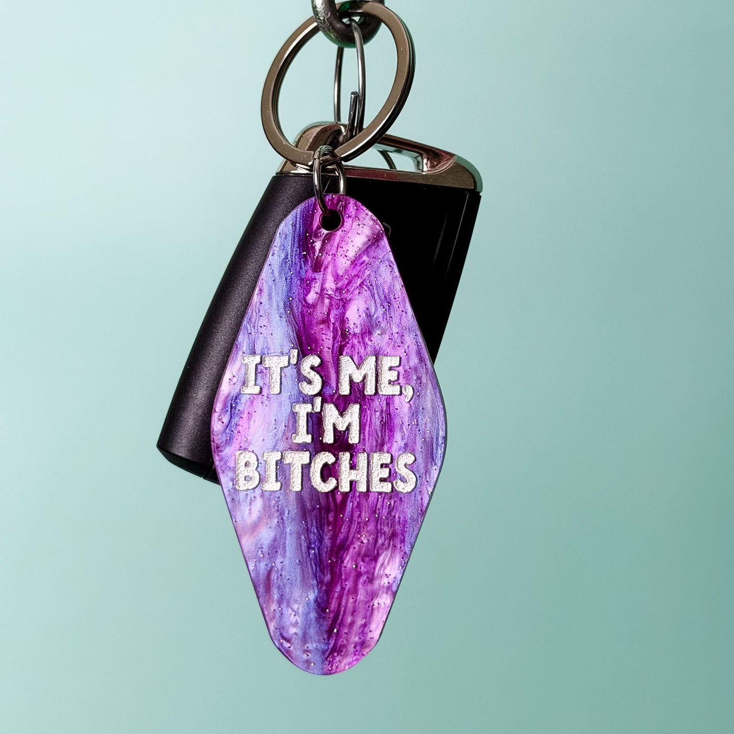 A keychain with a purple and pink marbled gemstone pendant and a black rectangular tag, hanging from a metal carabiner on a wooden rod against a light teal background. The pendant has white text that reads 'IT'S ME, I'M BITCHES'.