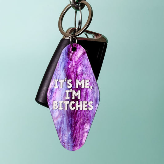 A keychain with a purple and pink marbled gemstone pendant and a black rectangular tag, hanging from a metal carabiner on a wooden rod against a light teal background. The pendant has white text that reads 'IT'S ME, I'M BITCHES'.