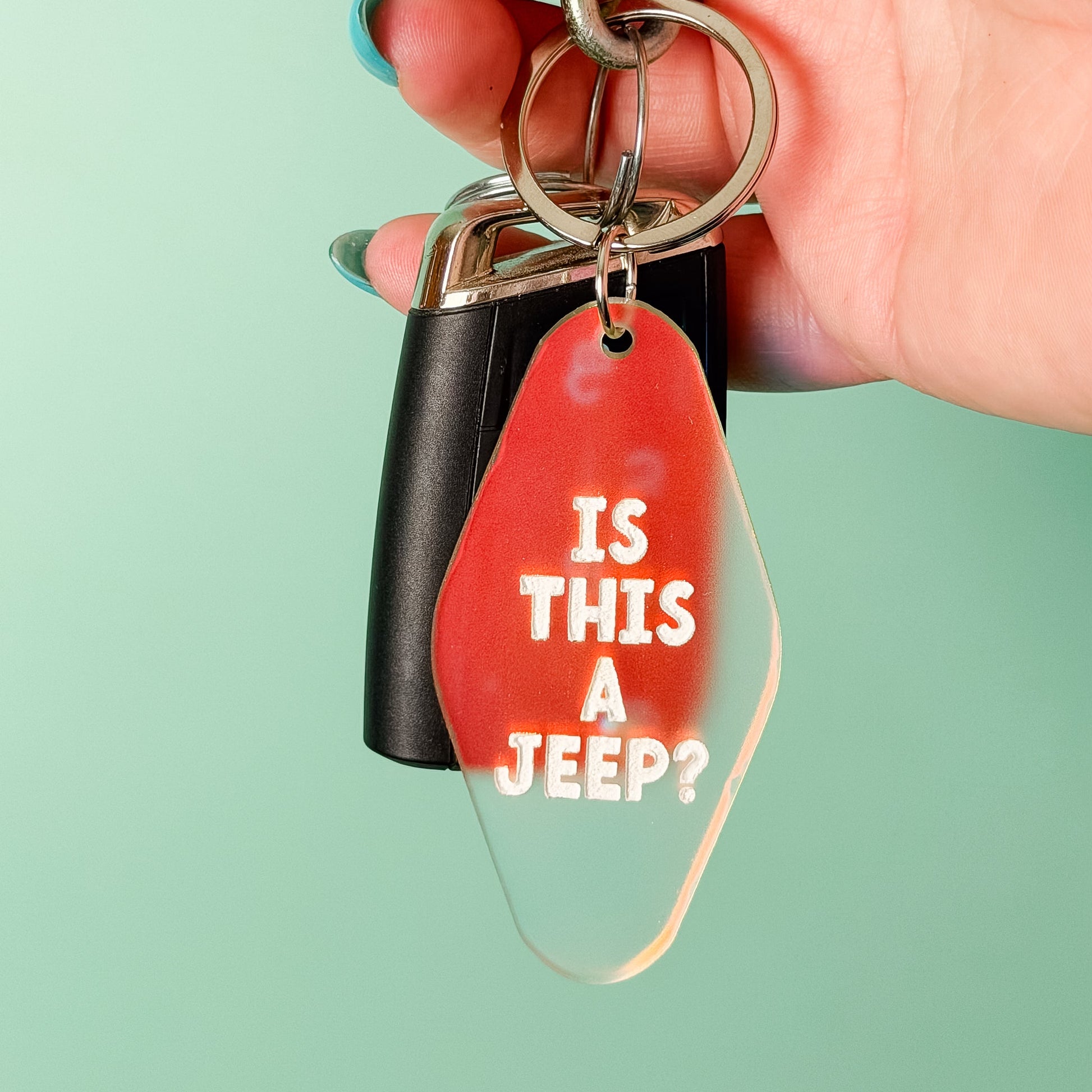 A hand with blue nail polish holds a keychain featuring a red translucent tag that reads 'IS THIS A JEEP?' in white text, alongside a black key fob and silver metal rings, against a solid mint green background.