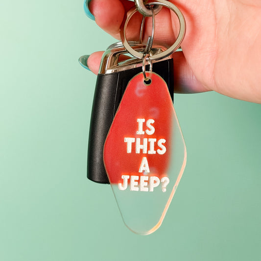 A hand with blue nail polish holds a keychain featuring a red translucent tag that reads 'IS THIS A JEEP?' in white text, alongside a black key fob and silver metal rings, against a solid mint green background.