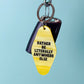 A yellow guitar-pick-shaped keychain with black text reading 'RATHER BE LITERALLY ANYWHERE ELSE' hangs from a silver carabiner and key ring, with a black key fob behind it, against a light blue background.