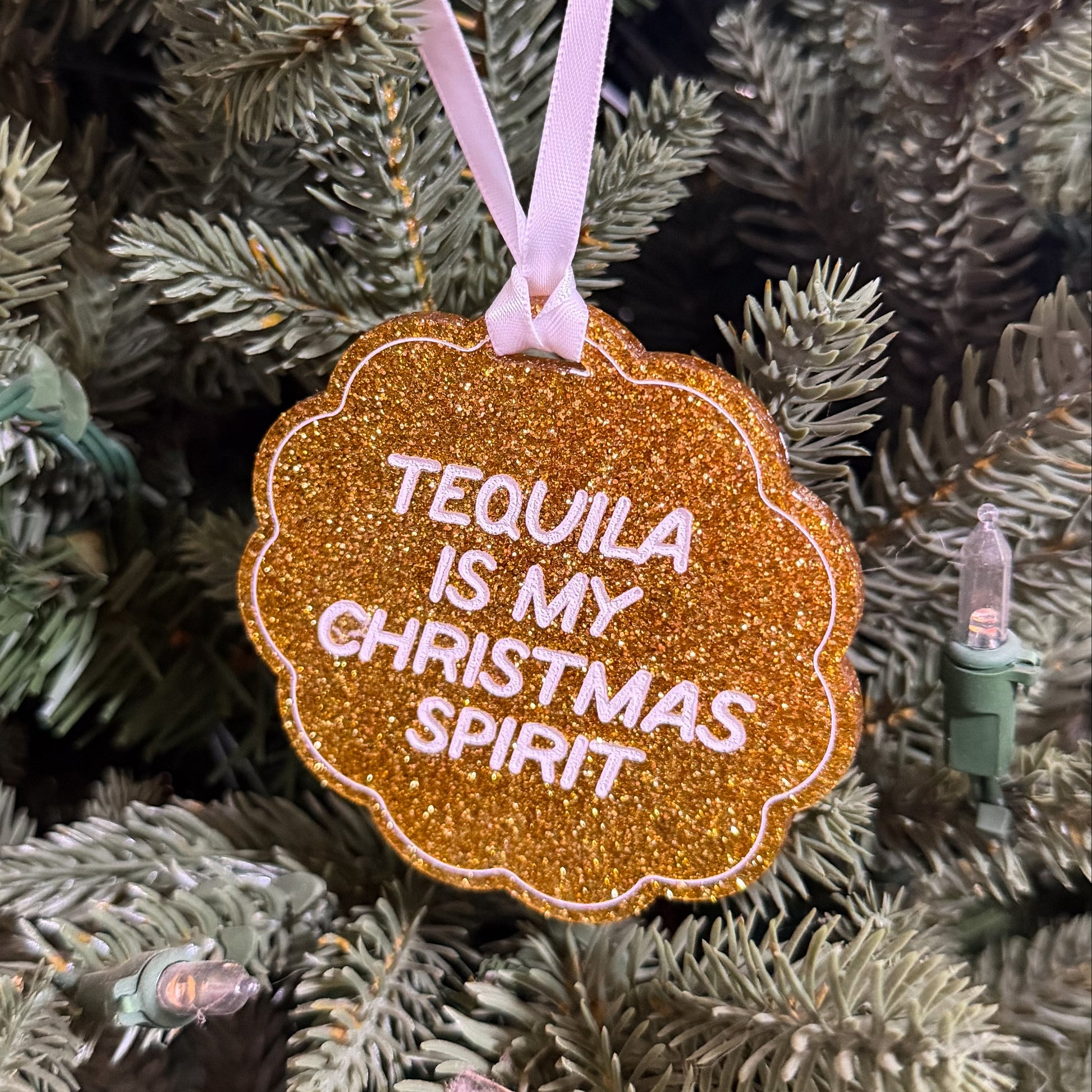 A scalloped glitter ornament in metallic copper-orange with white text reading 'TEQUILA IS MY CHRISTMAS SPIRIT,' hanging from a pale pink ribbon on a green Christmas tree.