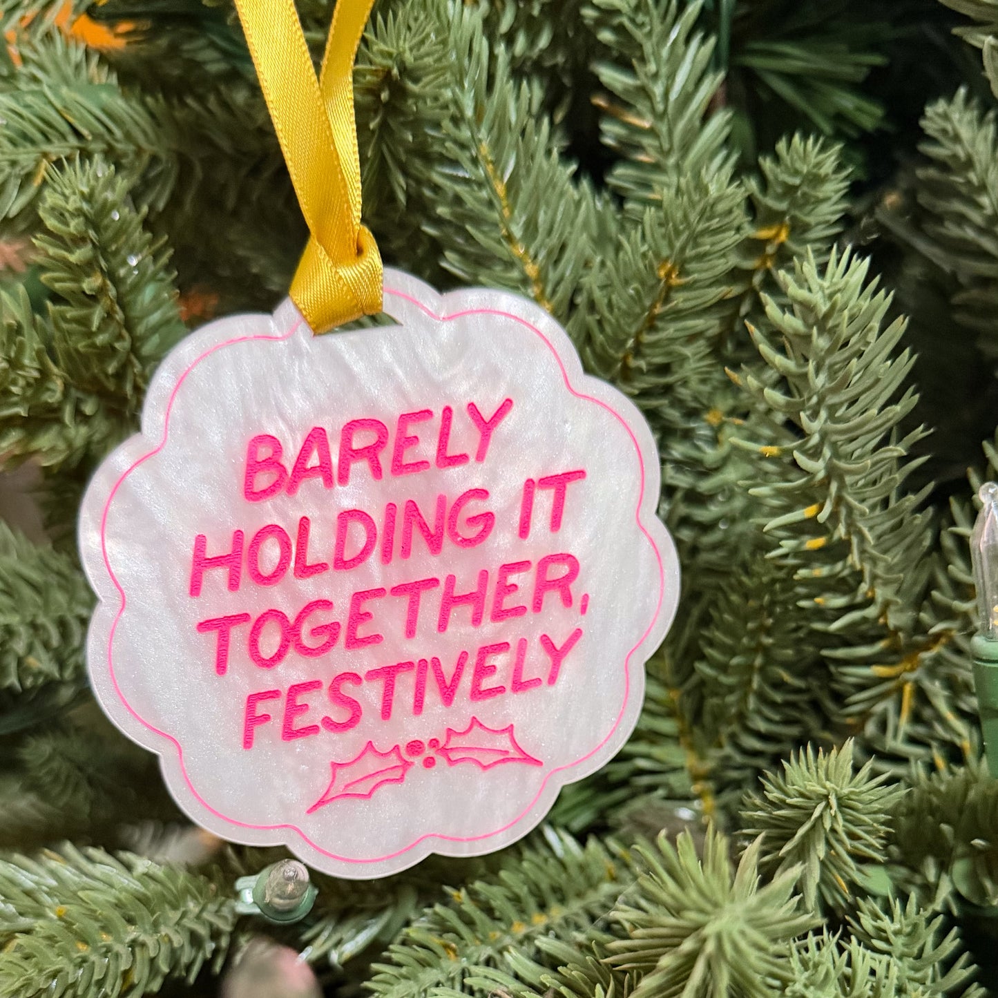 A scalloped white Christmas ornament with pink text reading 'BARELY HOLDING IT TOGETHER, FESTIVELY' and a pink outline, hanging from a yellow ribbon on green tree branches.