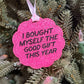 A bright pink glitter Christmas ornament with black text reading 'I BOUGHT MYSELF THE GOOD GIFT THIS YEAR' hanging from a white ribbon on a green Christmas tree.