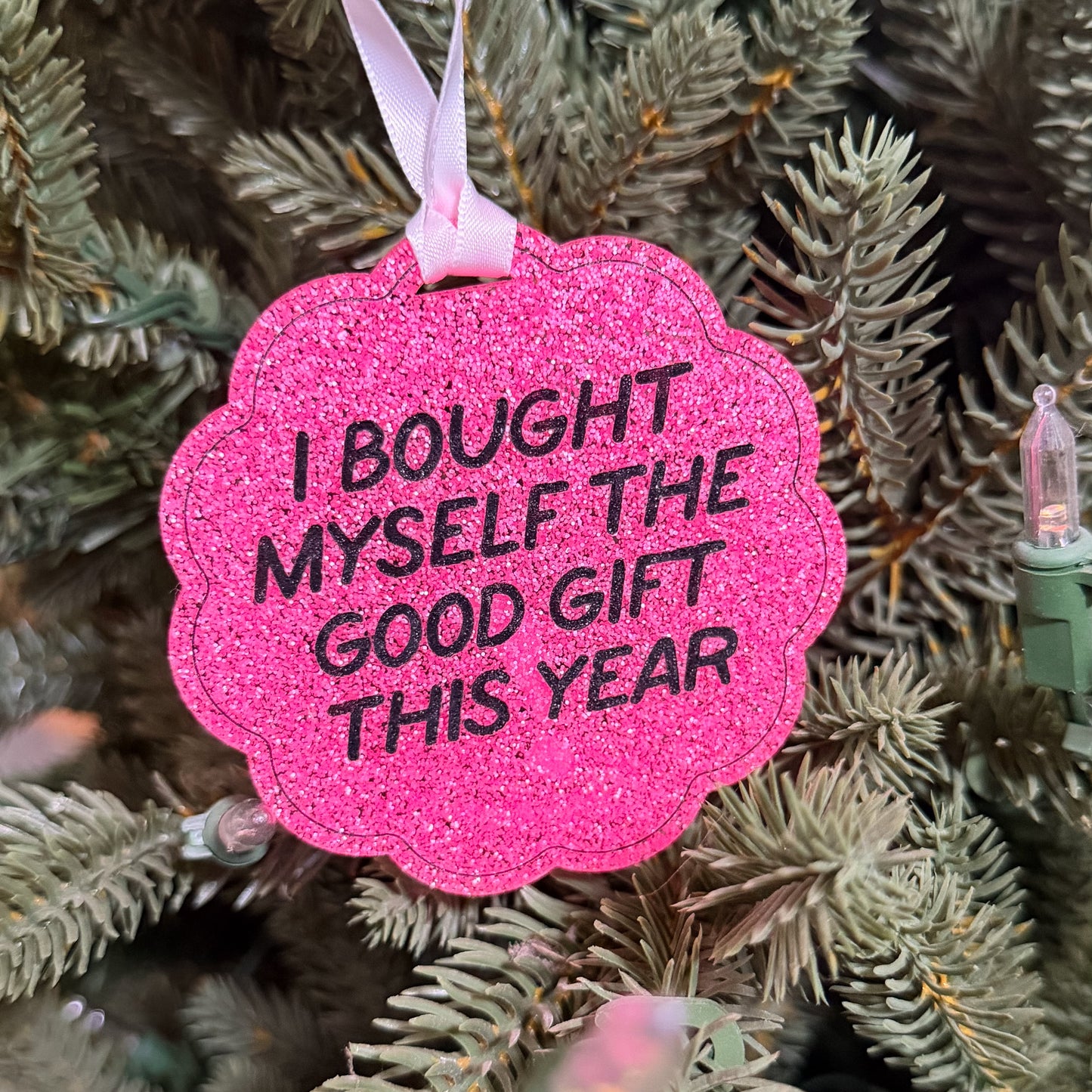 A bright pink glitter Christmas ornament with black text reading 'I BOUGHT MYSELF THE GOOD GIFT THIS YEAR' hanging from a white ribbon on a green Christmas tree.