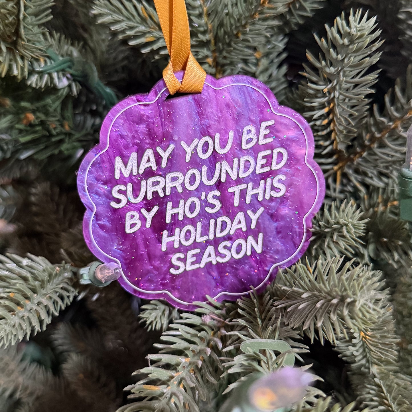 A purple scalloped Christmas ornament with white text reading 'MAY YOU BE SURROUNDED BY HO'S THIS HOLIDAY SEASON' hanging from an orange ribbon on a green pine tree.
