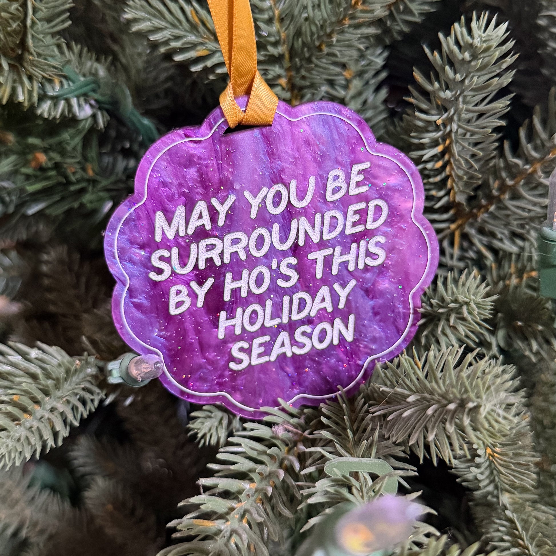 A purple scalloped Christmas ornament with white text reading 'MAY YOU BE SURROUNDED BY HO'S THIS HOLIDAY SEASON' hanging from an orange ribbon on a green pine tree.