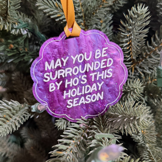A purple scalloped Christmas ornament with white text reading 'MAY YOU BE SURROUNDED BY HO'S THIS HOLIDAY SEASON' hanging from an orange ribbon on a green pine tree.