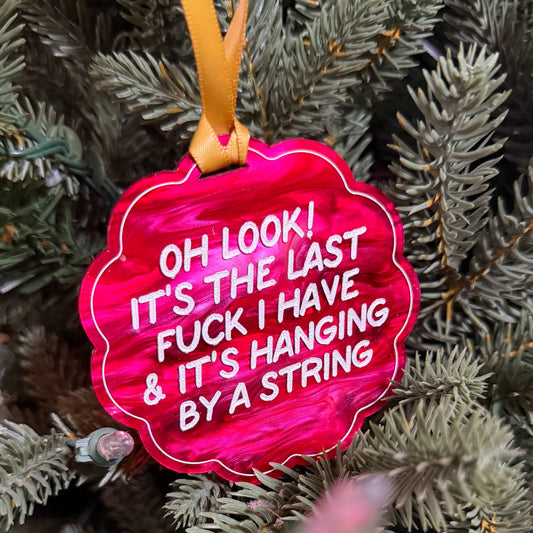 A bright pink scalloped Christmas ornament with white text hanging from a yellow ribbon on a green pine tree.