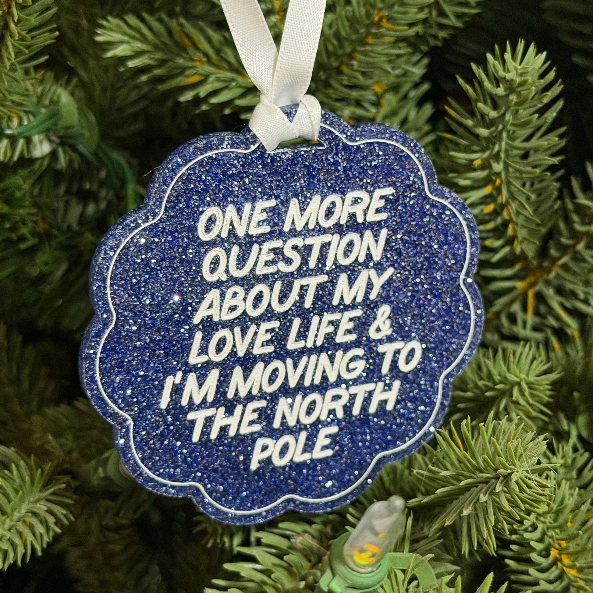 A scalloped blue glitter Christmas ornament with white text reading 'ONE MORE QUESTION ABOUT MY LOVE LIFE & I'M MOVING TO THE NORTH POLE', hanging from a white ribbon on a green pine tree.