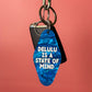 A blue marbled guitar-pick-shaped keychain with white text reading 'DELULU IS A STATE OF MIND' hangs from a silver carabiner on a wooden rod against a solid coral-pink background.
