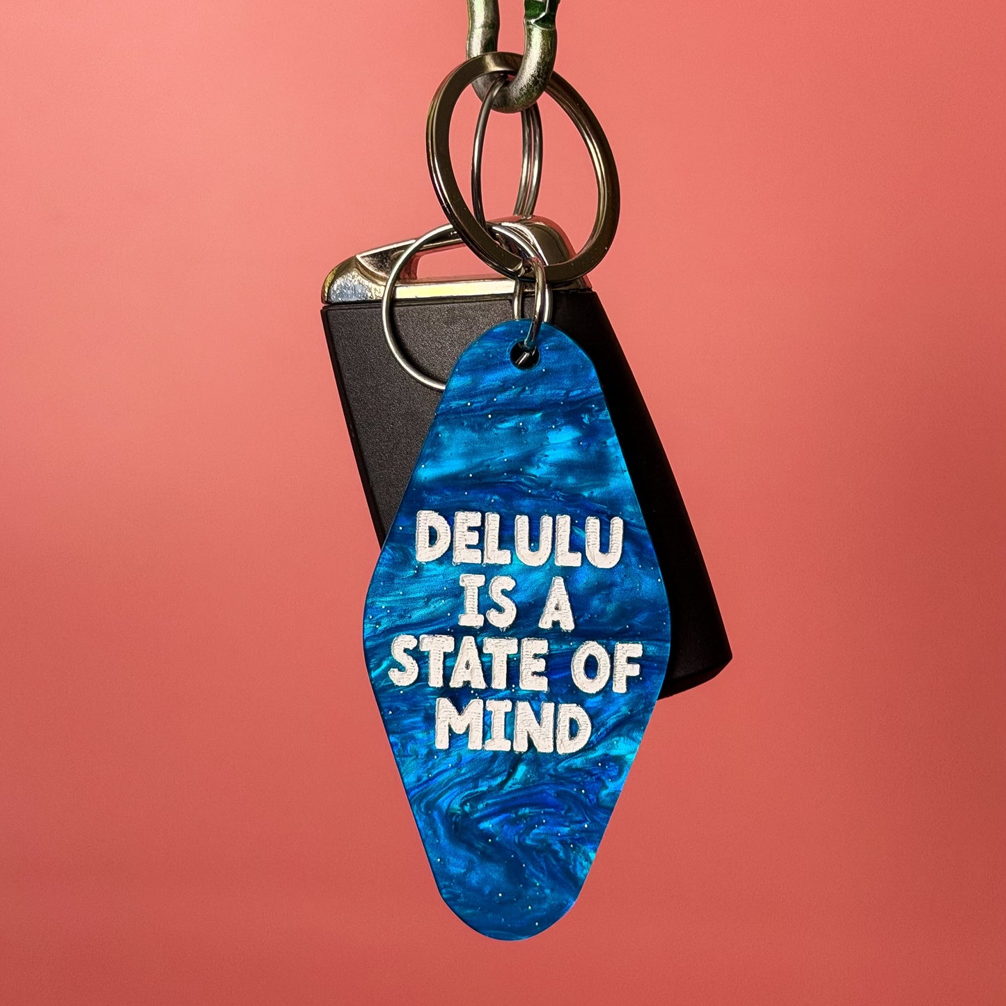 A blue marbled guitar-pick-shaped keychain with white text reading 'DELULU IS A STATE OF MIND' hangs from a silver carabiner on a wooden rod against a solid coral-pink background.