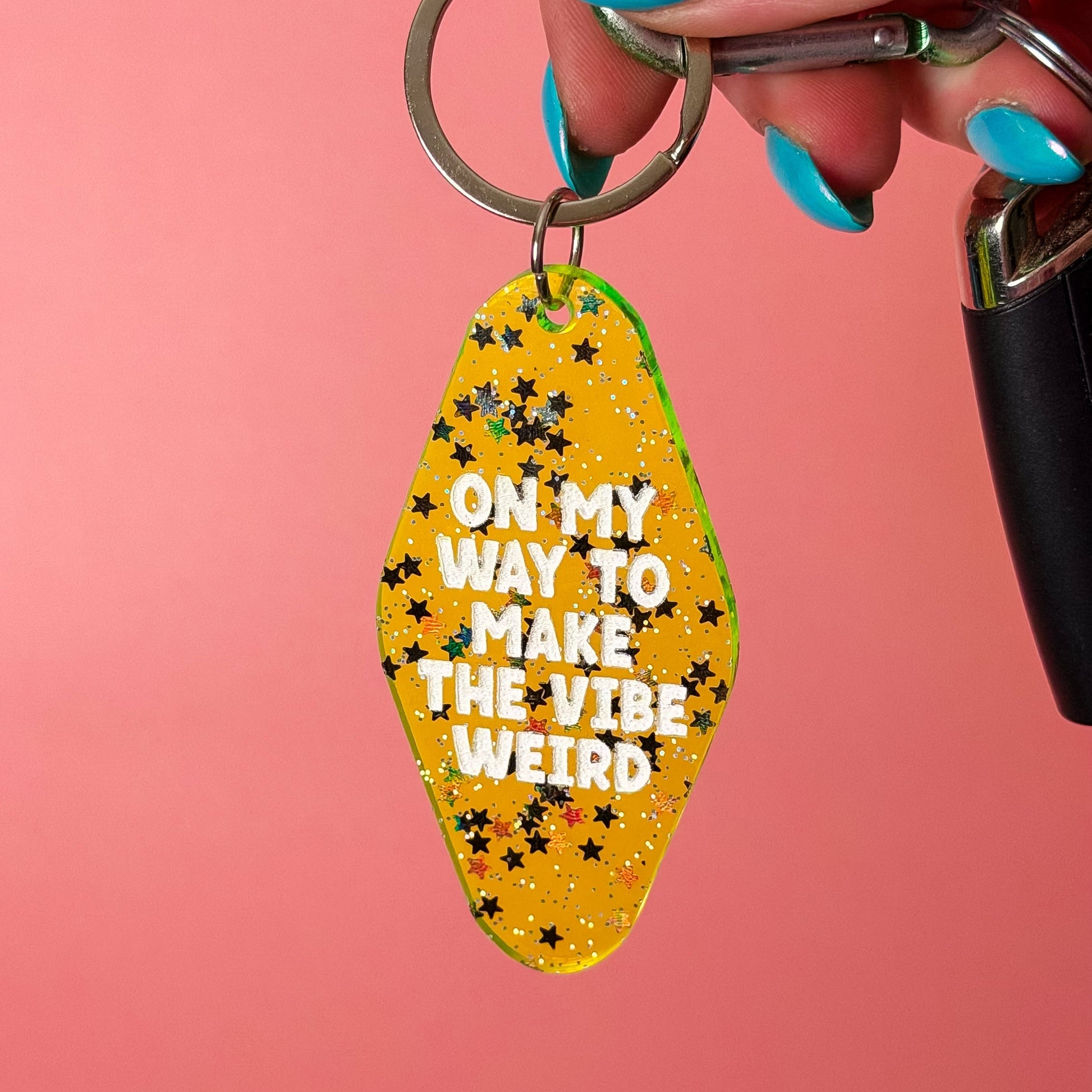 A hand with turquoise nail polish holds a yellow glitter keychain with black stars and the text 'ON MY WAY TO MAKE THE VIBE WEIRD' against a solid pink background.