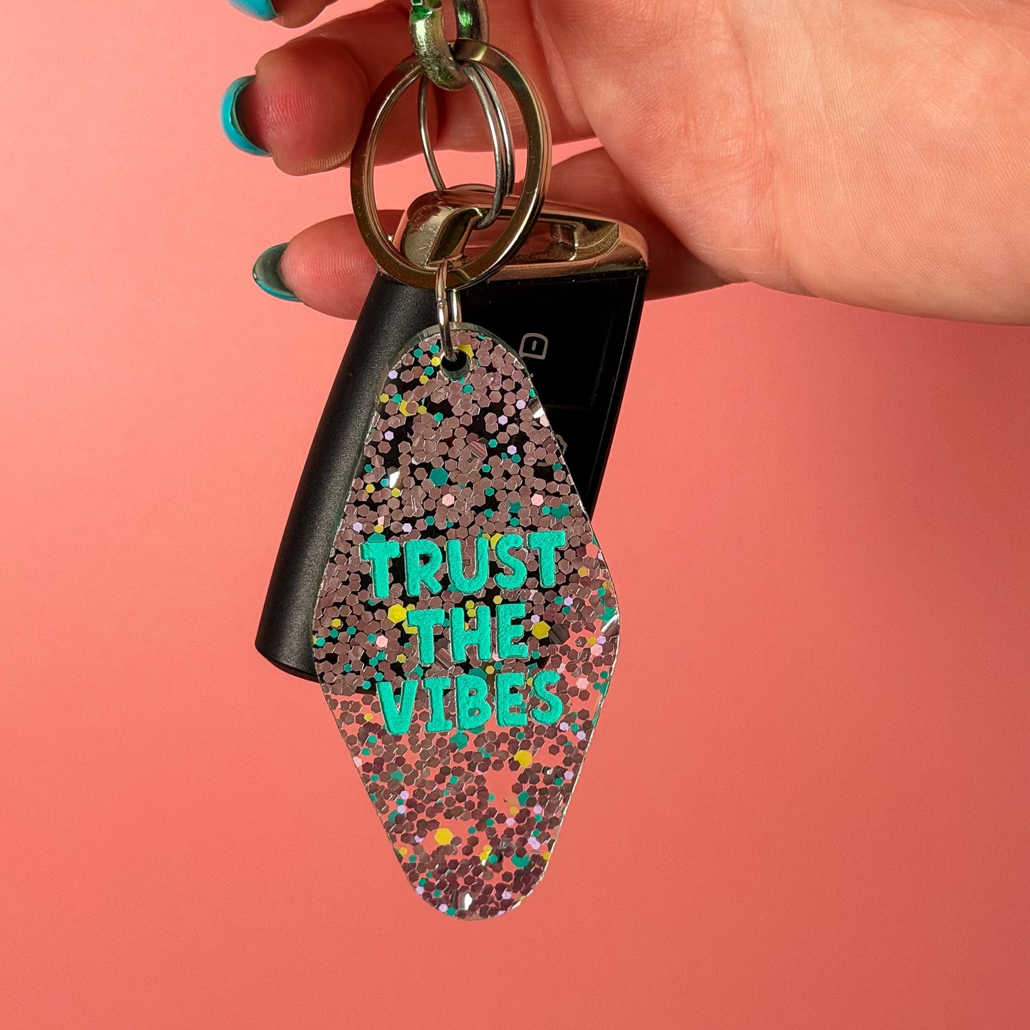 A hand with turquoise nail polish holds a keychain featuring a glittery, diamond-shaped charm with the text 'TRUST THE VIBES' in bright teal. The keychain also includes a black car key fob and a green carabiner, set against a solid coral-pink background.